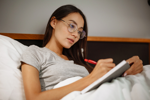 woman laying in bed while writing in a journal with a red pen